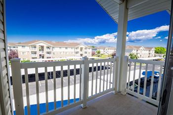 A balcony with a white railing overlooks a parking lot and apartment buildings.